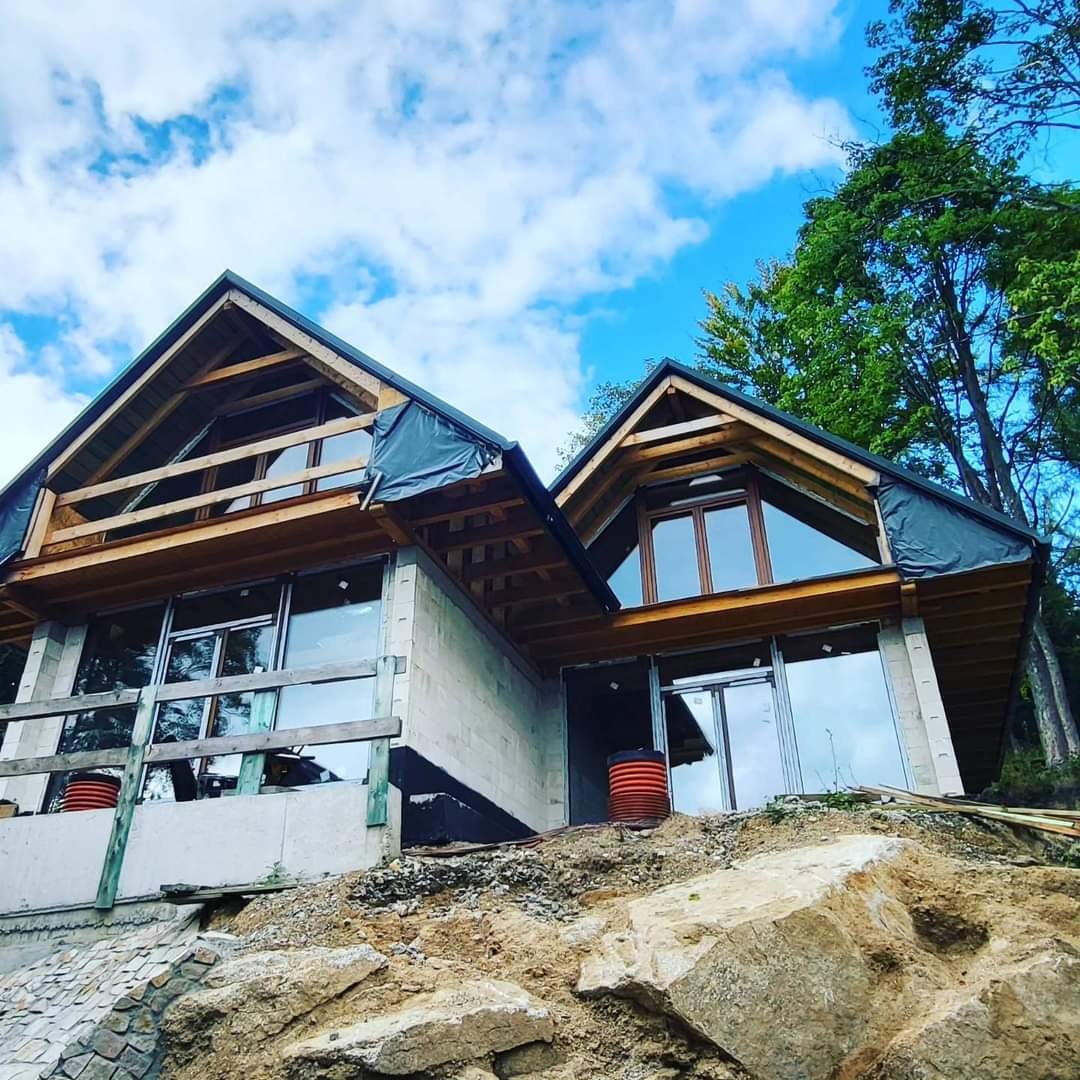 Dom w stanie surowym otwartym na zboczu wzgórza, widoczne duże przeszklenia i drewniana konstrukcja dachu, zabezpieczona folią, dwie beczki na pierwszym planie.