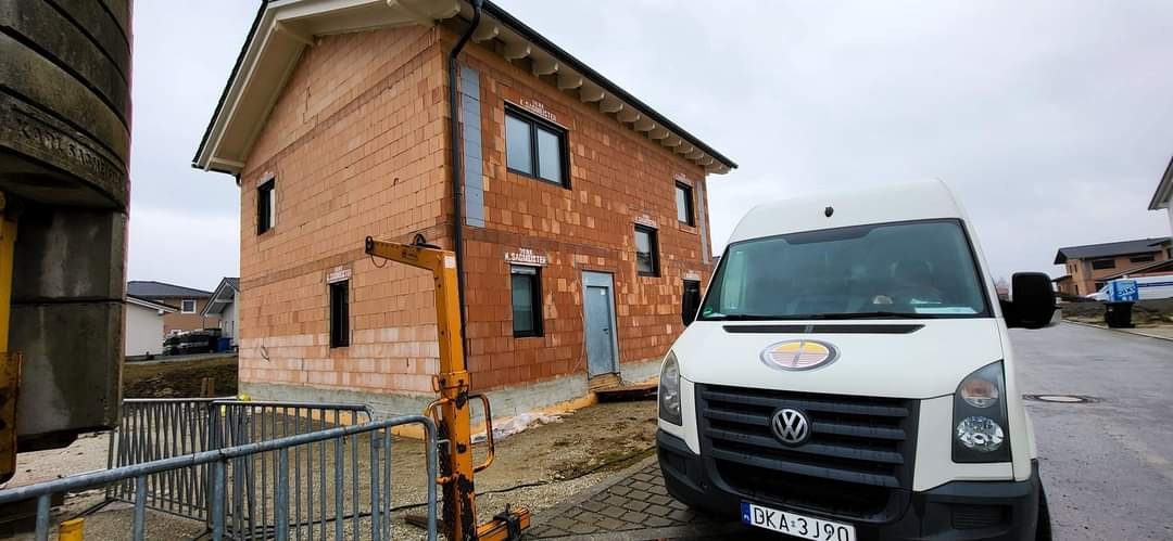 Montaż stolarki okiennej w surowym budynku z cegły, widoczny podnośnik i samochód dostawczy firmy.