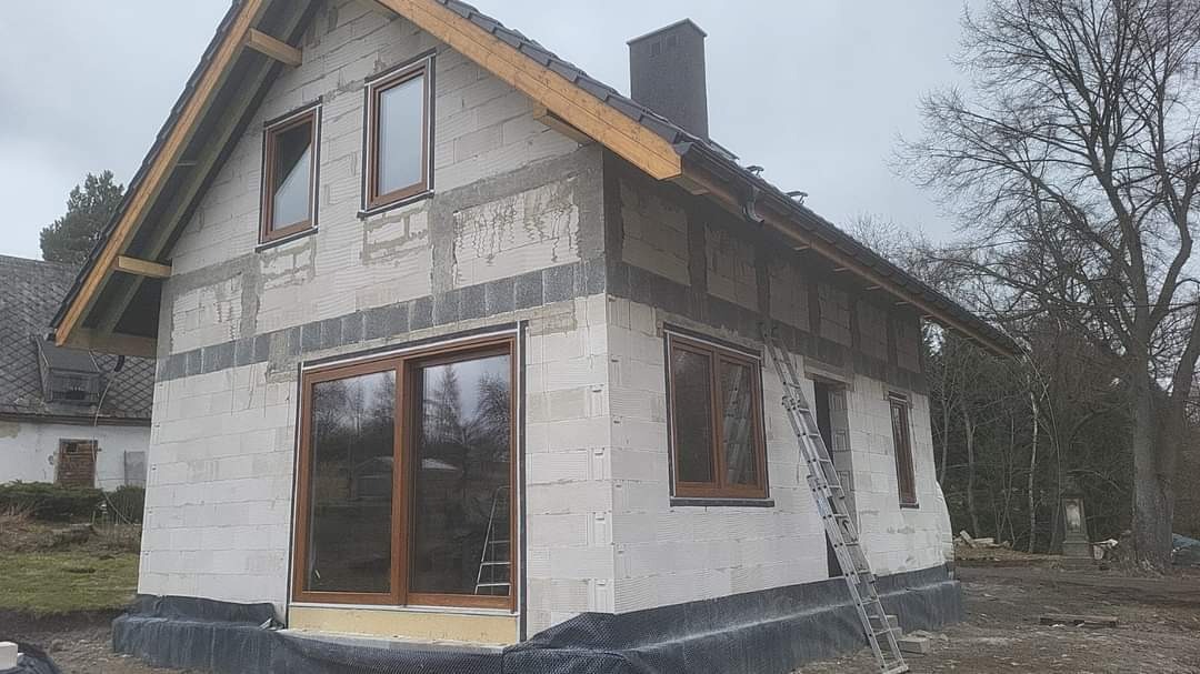 Surowy stan domu w trakcie budowy z zamontowanymi oknami w drewnianych ramach, widoczna drabina oparta o ścianę i fragment dachu.