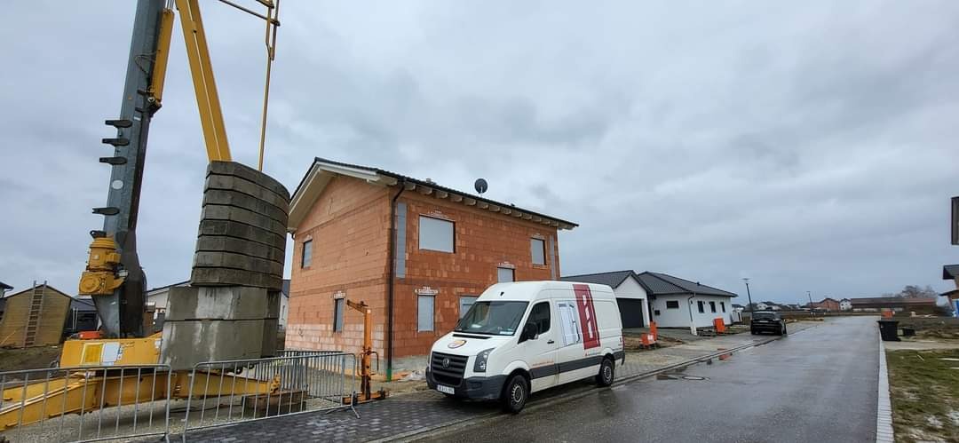 Budowa domu jednorodzinnego z czerwonej cegły, widoczny dźwig z obciążnikami, oraz biały samochód dostawczy firmy budowlanej na tle pochmurnego nieba.