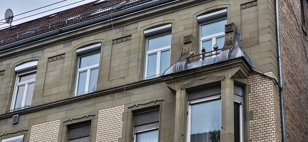 Fasada kamienicy z oknami i częściowo opuszczonymi roletami zewnętrznymi, widoczne dekoracyjne elementy architektoniczne i ceglana ściana boczna.