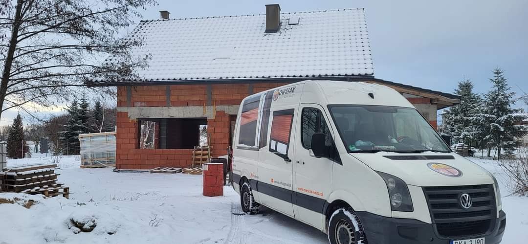 Biały samochód dostawczy firmy na tle domu w trakcie budowy, z widocznymi otworami okiennymi i śniegiem na dachu, w zimowej scenerii.