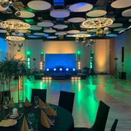 Sala bankietowa przygotowana na imprezę, z oświetleniem scenicznym w odcieniach zieleni i niebieskiego, dekoracjami sufitowymi w postaci dyskotekowych kul i okrągłych lamp, oraz stolami nakrytymi...