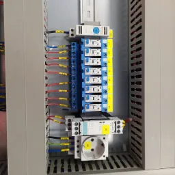 Wnętrze szafy sterowniczej z widocznymi przekaźnikami Finder, modułami Siemens Sirius, okablowaniem oznaczonym żółtymi etykietami i gniazdem elektrycznym. Uporządkowany układ komponentów.