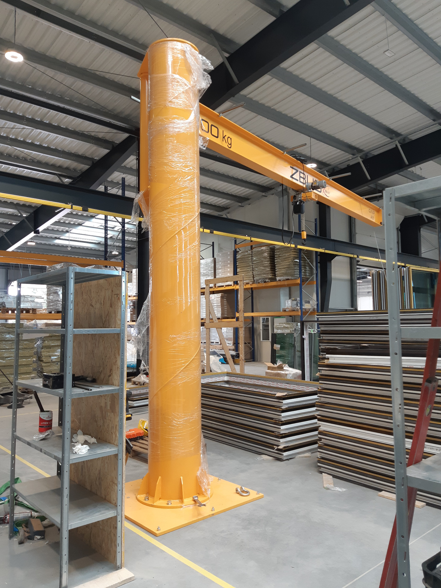 Nowa, żółta kolumna dźwigu obrotowego owinięta folią ochronną, stojąca w hali produkcyjnej przed regałami z oknami i ramami okiennymi.