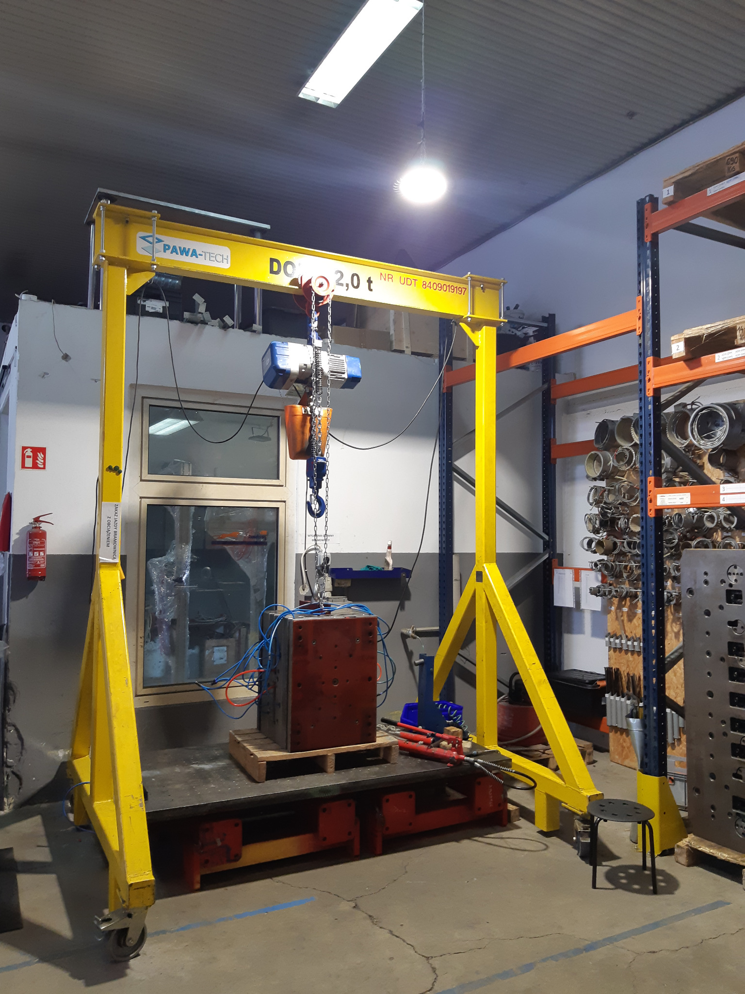 Żółty suwnicowy dźwig bramowy PAWA-TECH o udźwigu 2,0 tony, podnoszący metalowy blok na palecie w warsztacie, z widocznymi przewodami pneumatycznymi i elementami wyposażenia.