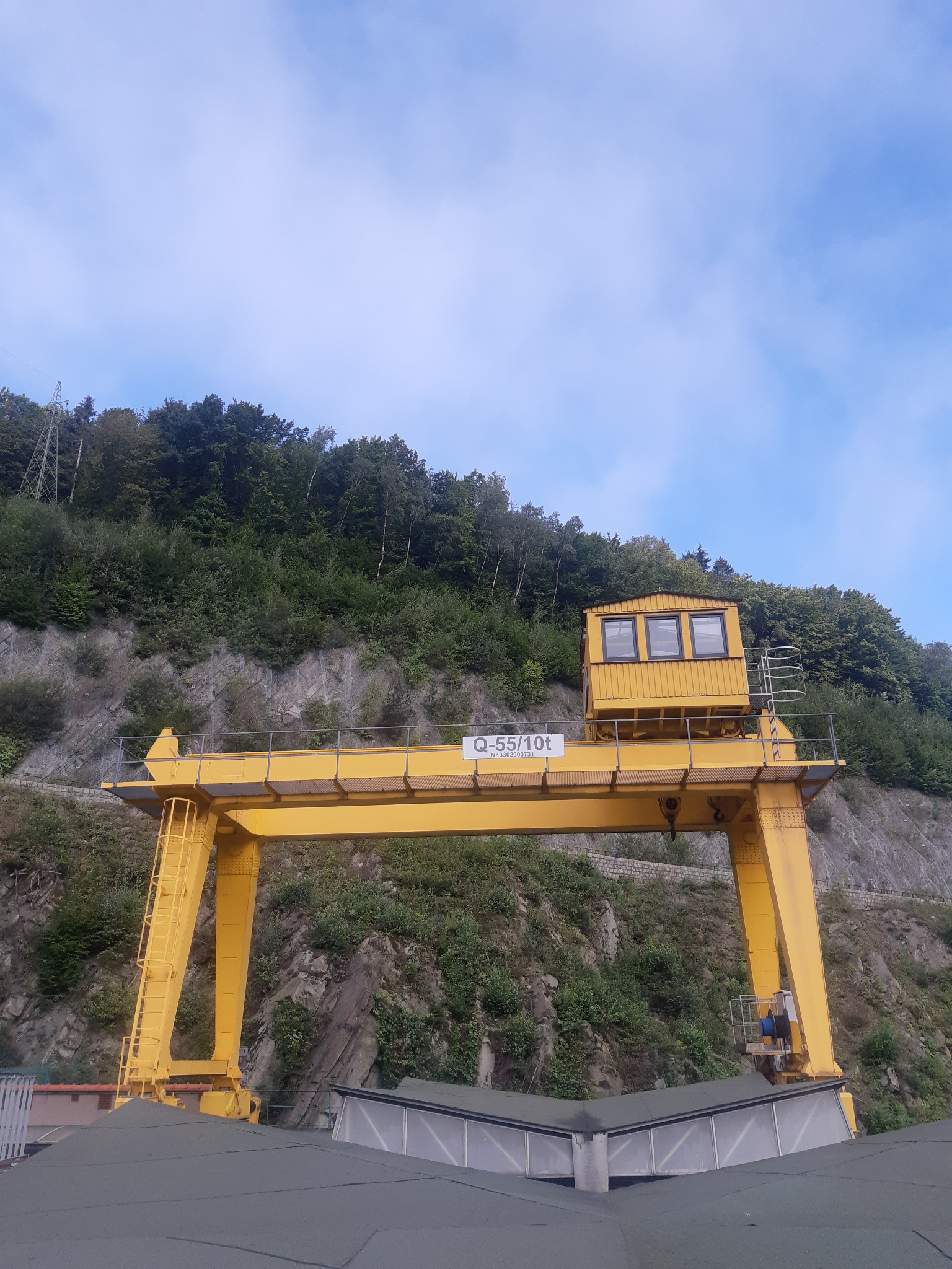 Żółty suwnicowy dźwig bramowy Q-55/10t z kabiną operatora, na tle zalesionego zbocza i błękitnego nieba.