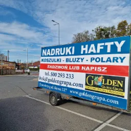 Reklamowy baner na przyczepce w Tarnobrzegu, informujący o nadrukach, haftach na koszulkach, bluzach i polarach, z numerem telefonu i adresem strony internetowej firmy Golden Global Grupa.