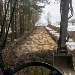 Widok z kabiny koparki na przygotowany teren pod drogę, z widocznymi śladami gąsienic i resztkami śniegu na obrzeżach, otoczony lasem.