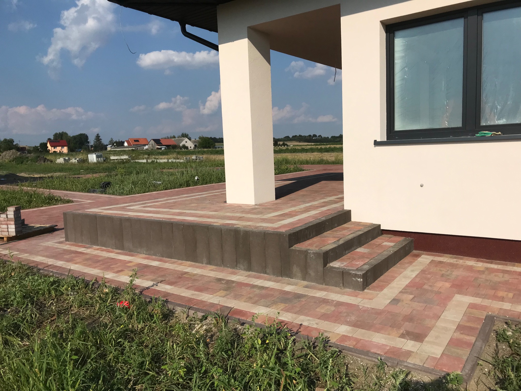 Wejście do domu z kostki brukowej w odcieniach czerwieni i beżu, schody z ciemnoszarych bloków, w tle widok na okoliczne domy i błękitne niebo.