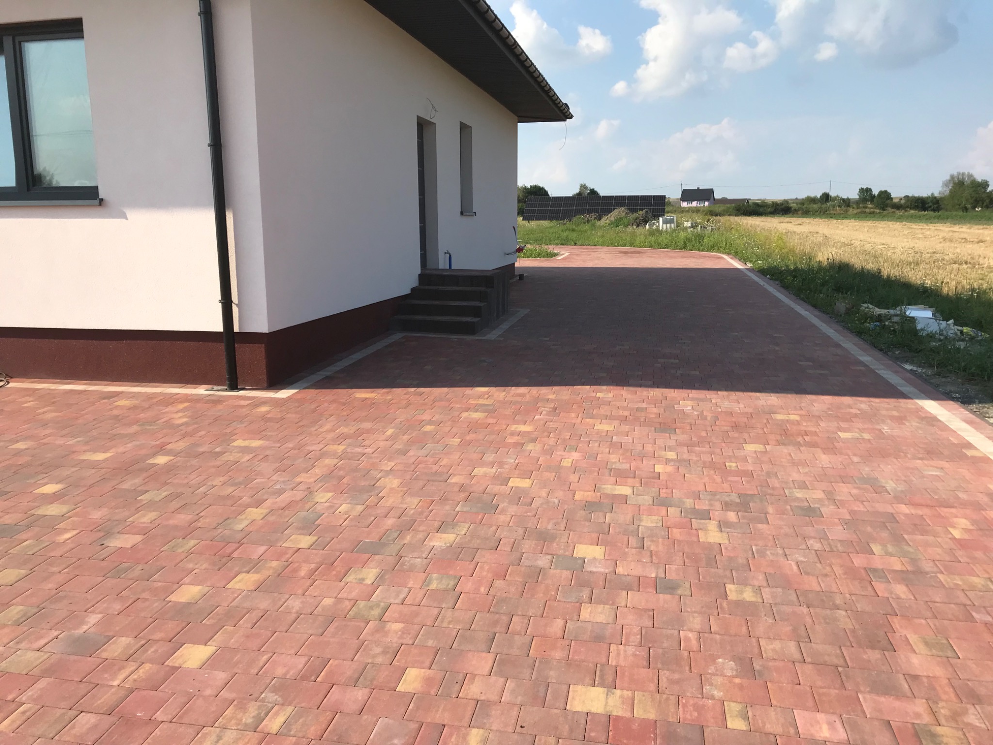 Nowo ułożony podjazd z kostki w odcieniach czerwieni i żółci przy białym domu z brązowymi akcentami, widok na pole w tle.