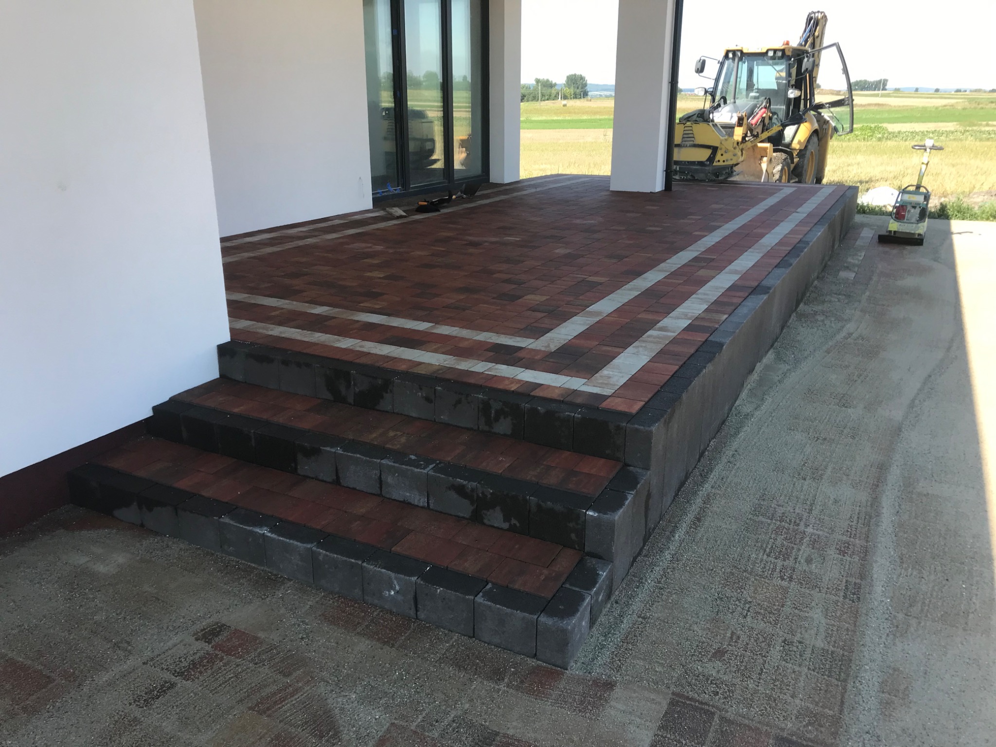 Ułożony taras z kostki brukowej z trzema schodami, obrzeżem z szarej kostki, z widocznym traktorem i ubijarką w tle.