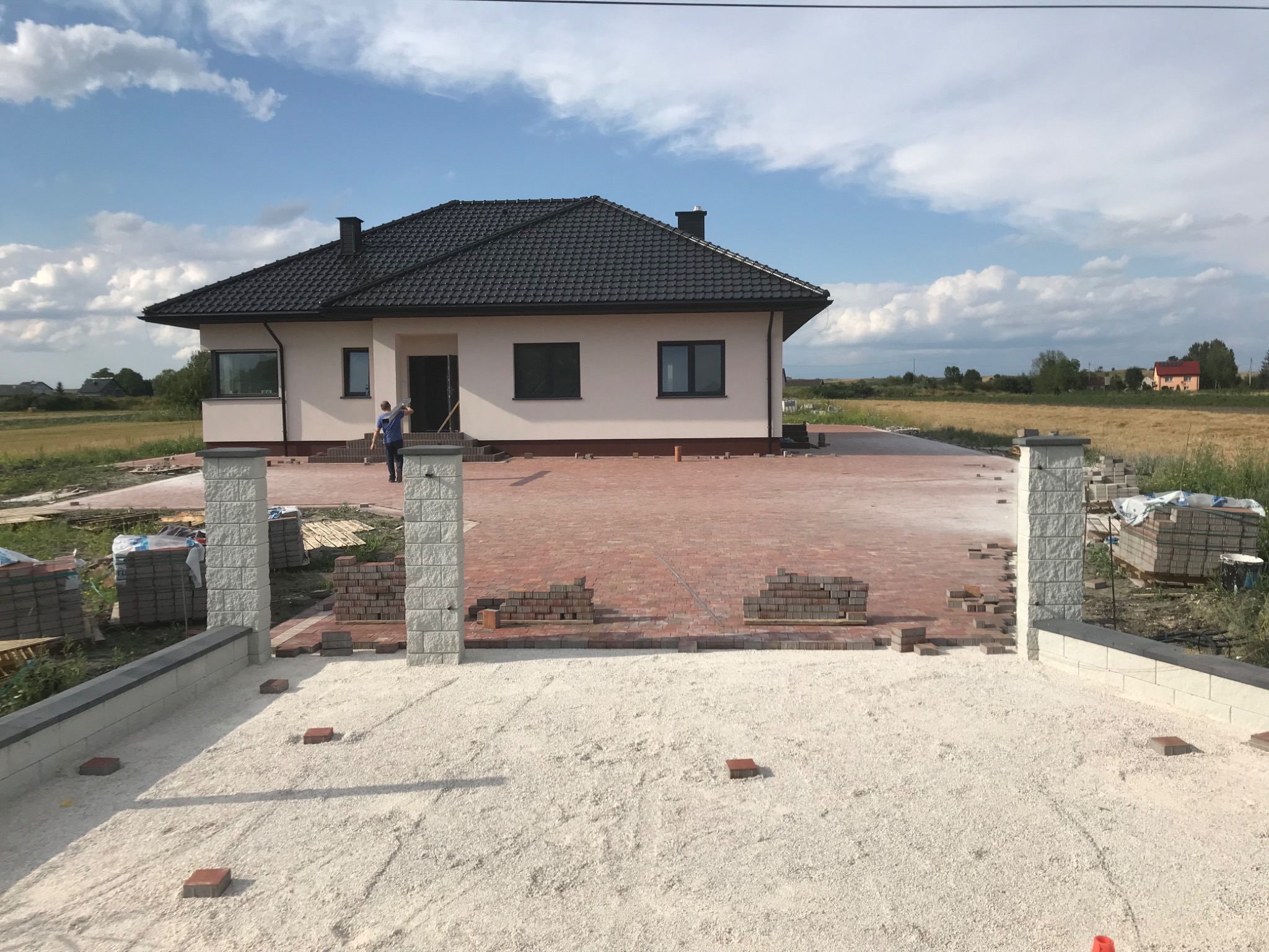 W trakcie układania czerwonej kostki brukowej na podjeździe przed domem jednorodzinnym. Widoczne przygotowane podłoże z piasku, palety z kostką i fragmenty ułożonej nawierzchni.