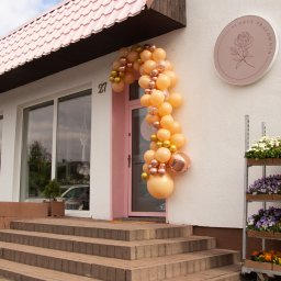 Flores Pracownia Bierzglinek - Kwiaciarnia Września - Wejście do kwiaciarni ozdobione girlandą z beżowych i złotych balonów, obok regał z kolorowymi kwiatami w doniczkach, szyld z logo firmy.