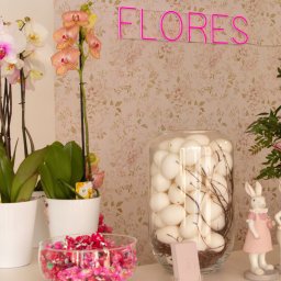 Flores Pracownia Bierzglinek - Kwiaciarnia Września - Aranżacja wnętrza kwiaciarni z różnokolorowymi storczykami w białych doniczkach, szklanym wazonem z białymi jajkami, miseczką cukierków, bukietem kwiatów i dekoracyjnymi króliczkami na tle ściany...