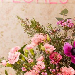 Flores Pracownia Bierzglinek - Kwiaciarnia Września - Delikatny bukiet z różowych goździków, róż i anemonów na tle ściany z napisem 'FLORES' wykonanym z różowych neonowych rurek.