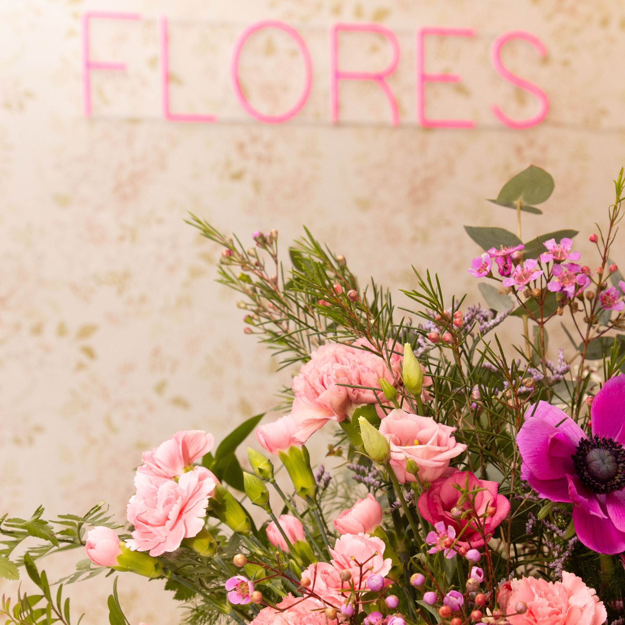 Kompozycja kwiatowa z różowych goździków, eustomy, anemonu i drobnych fioletowych kwiatów na tle ściany z napisem 'FLORES' wykonanym z różowych rurek.