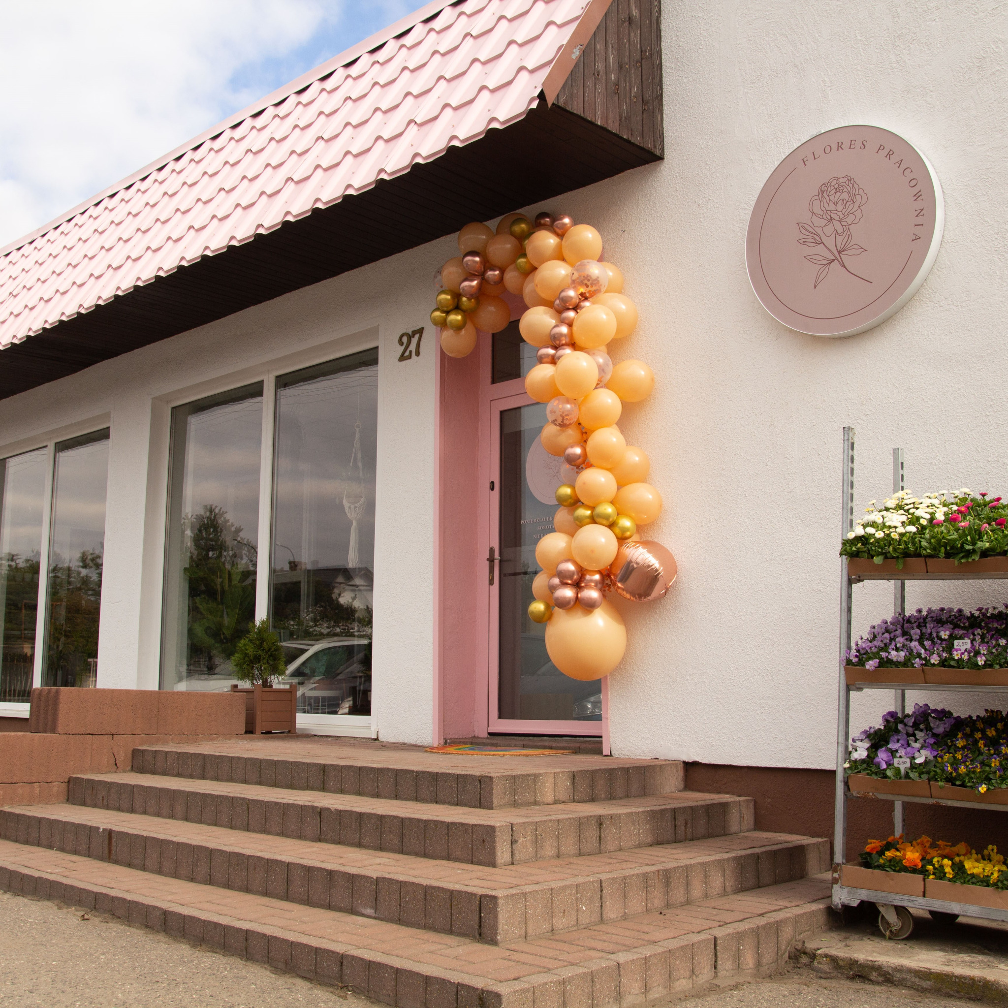Wejście do kwiaciarni ozdobione girlandą z brzoskwiniowych i złotych balonów, z boku regał z kwitnącymi bratkami i stokrotkami, nad drzwiami szyld z logo kwiaciarni.