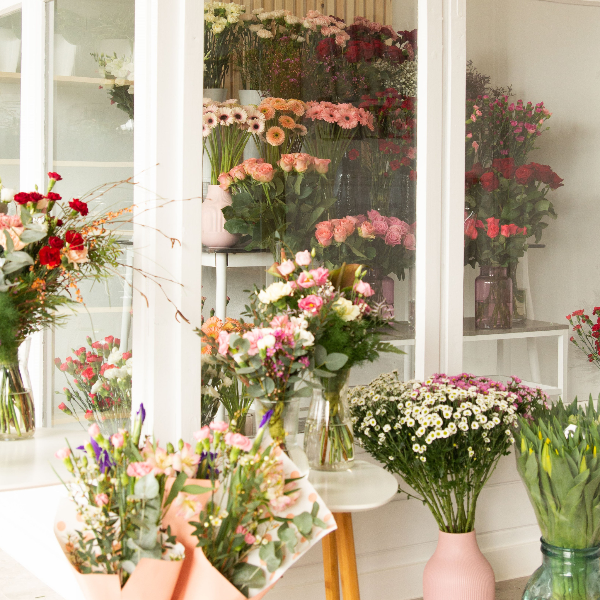 Witryna kwiaciarni z różnorodnymi bukietami kwiatów w wazonach, widoczne gerbery, róże i inne kwiaty cięte, część bukietów zapakowana w ozdobny papier.