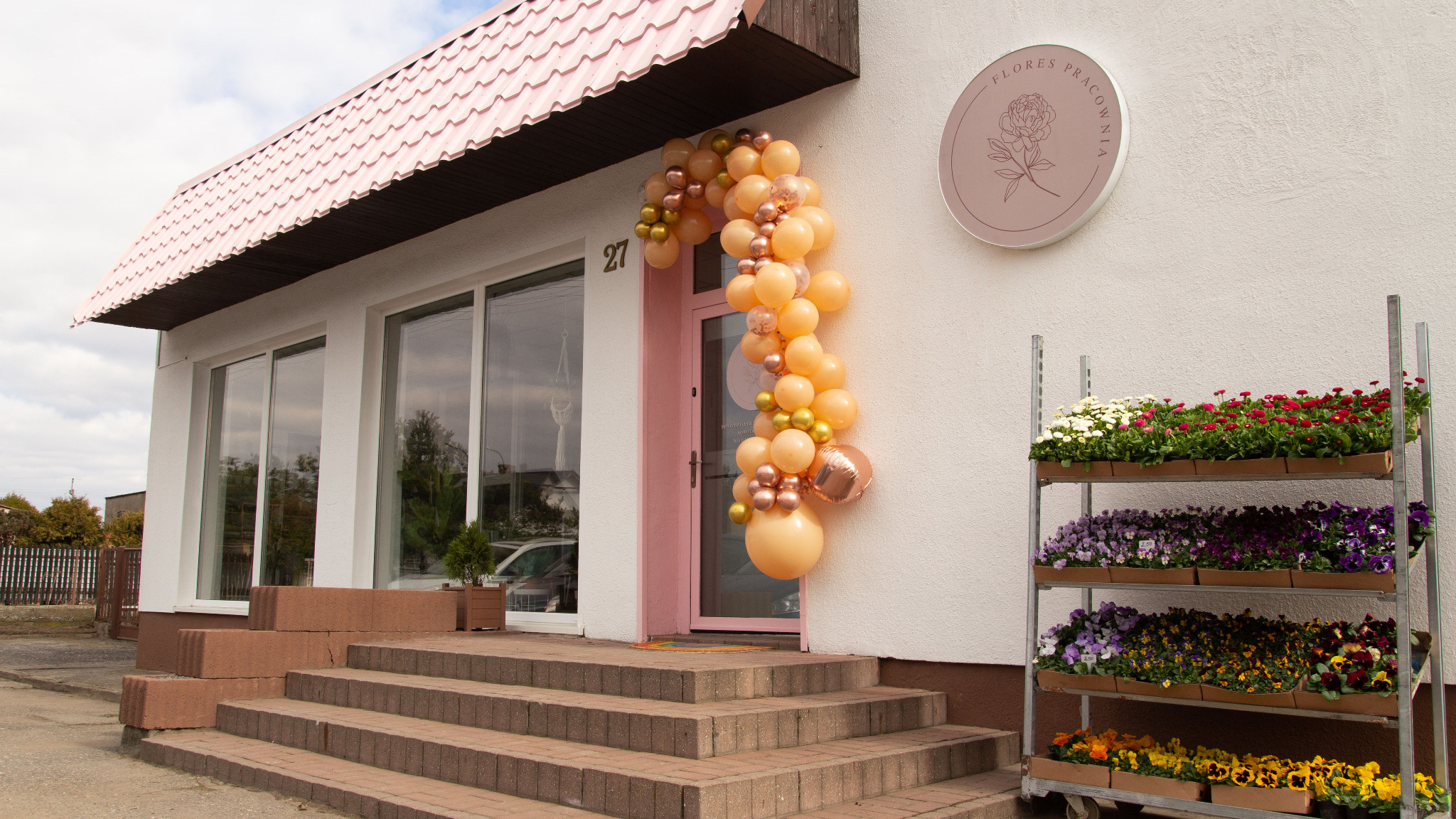 Wejście do kwiaciarni ozdobione girlandą z beżowych i złotych balonów, obok regał z kolorowymi kwiatami w doniczkach, szyld z logo firmy.