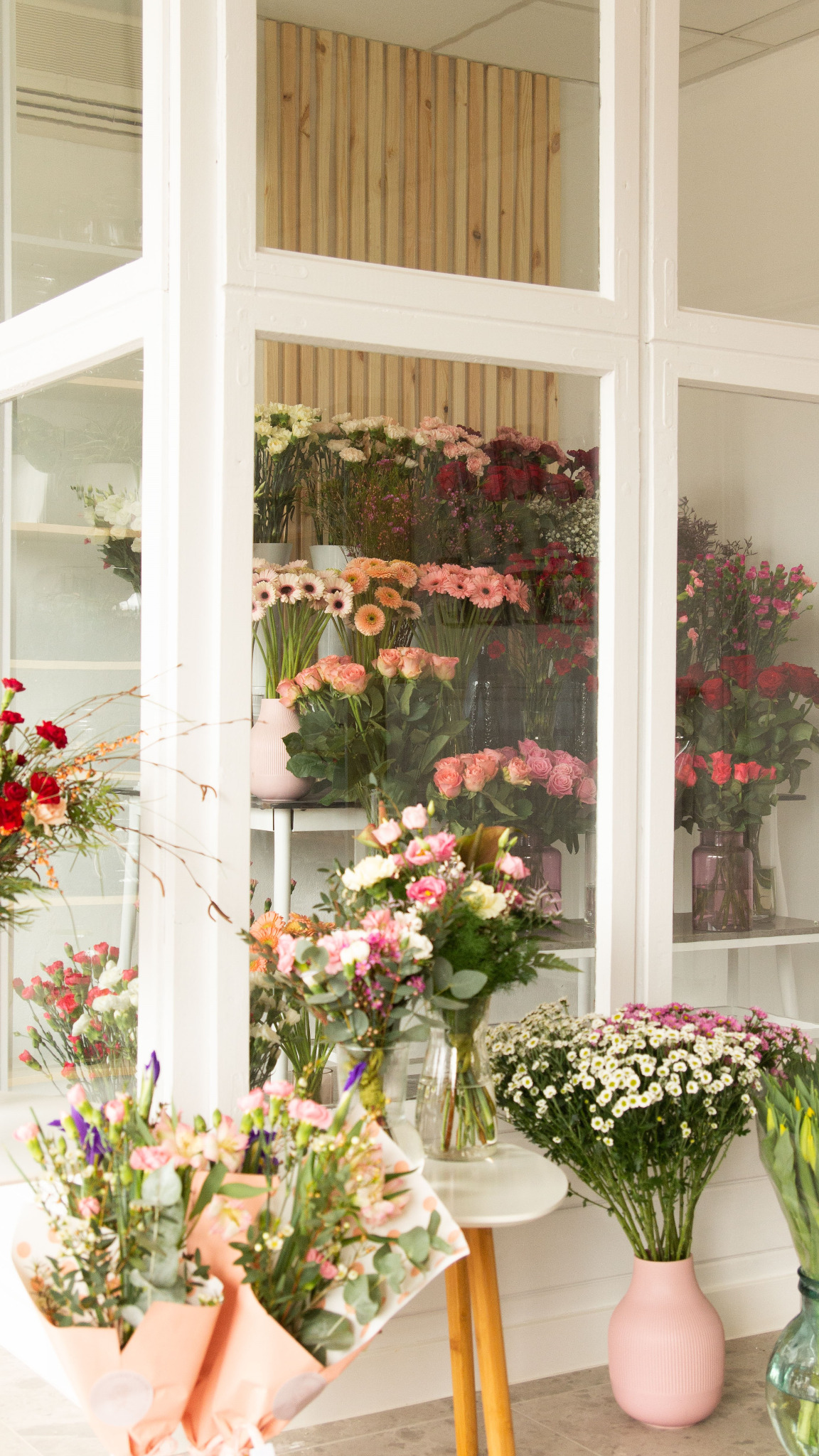 Kwiaty w wazonach i bukiety na wystawie kwiaciarni, widoczne przez białe ramy okienne.