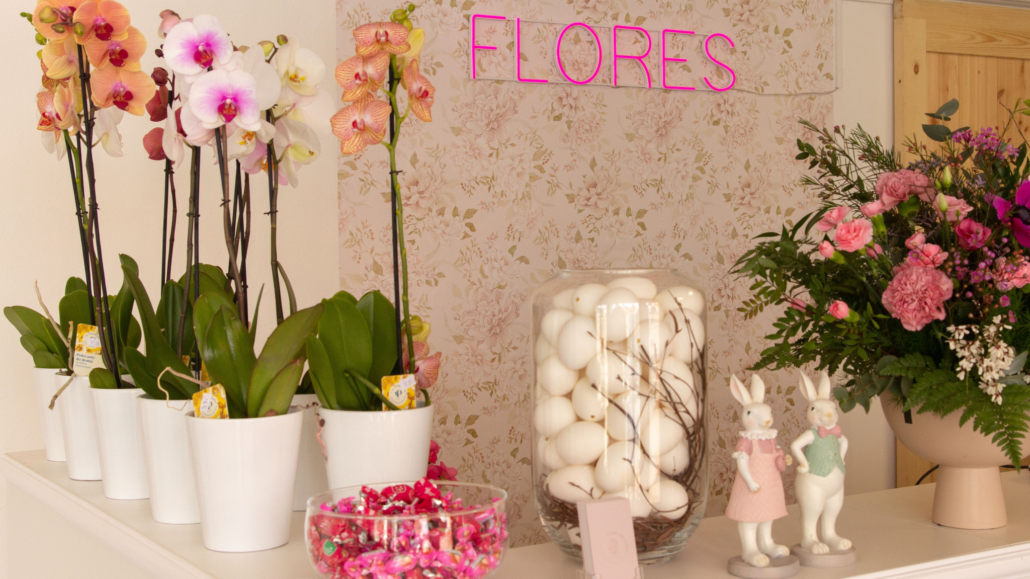 Aranżacja wnętrza kwiaciarni z różnokolorowymi storczykami w białych doniczkach, szklanym wazonem z białymi jajkami, miseczką cukierków, bukietem kwiatów i dekoracyjnymi króliczkami na tle ściany...