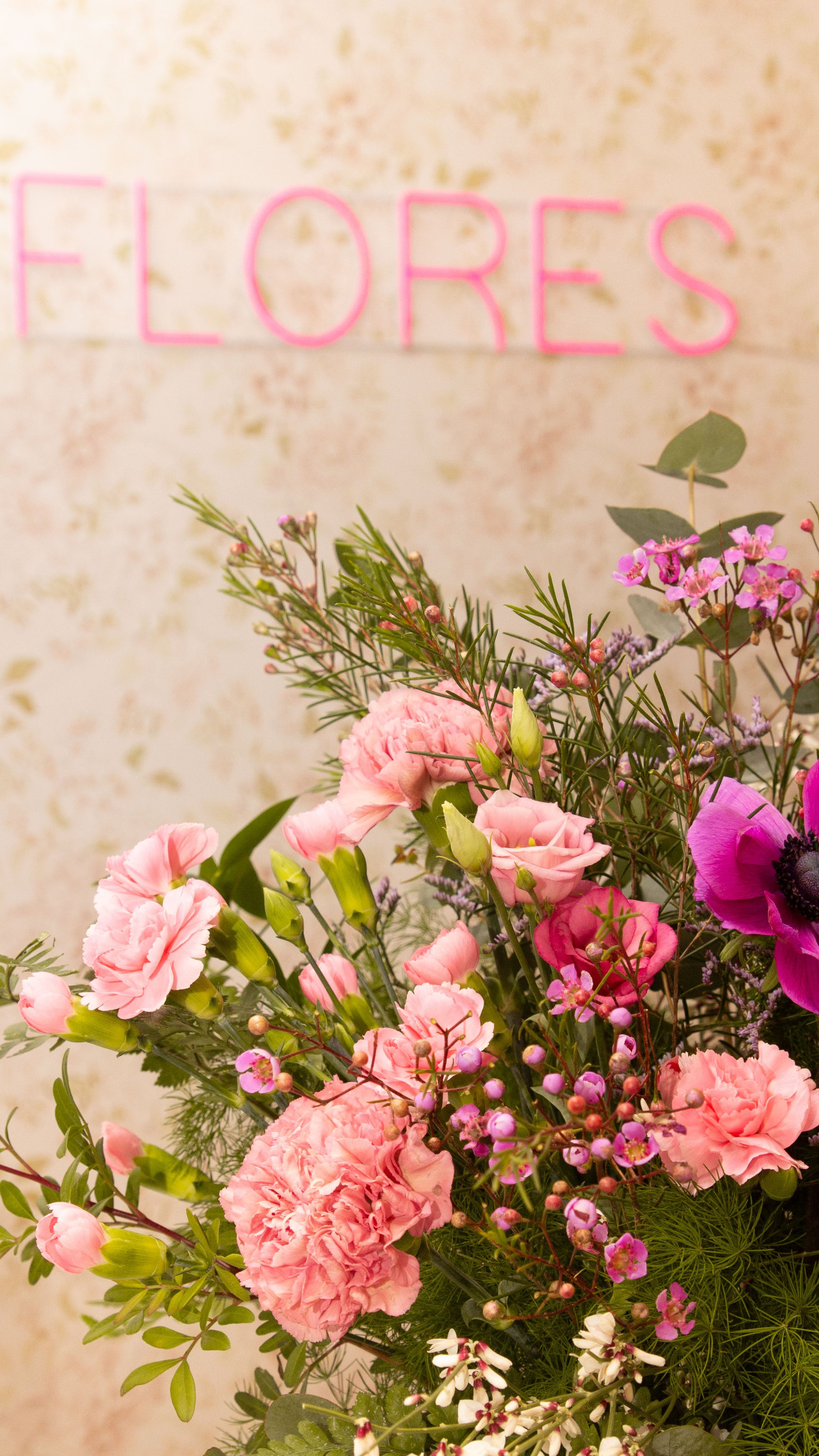 Delikatny bukiet z różowych goździków, róż i anemonów na tle ściany z napisem 'FLORES' wykonanym z różowych neonowych rurek.