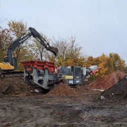 Żółta koparka Volvo załadowuje gruz do mobilnego przesiewacza Sandvik QJ341 na placu budowy z drzewami w tle.