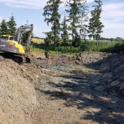 Żółto-szara koparka Volvo pracuje przy wykopie długiego rowu w ziemi, widok na teren budowy z drzewami w tle.