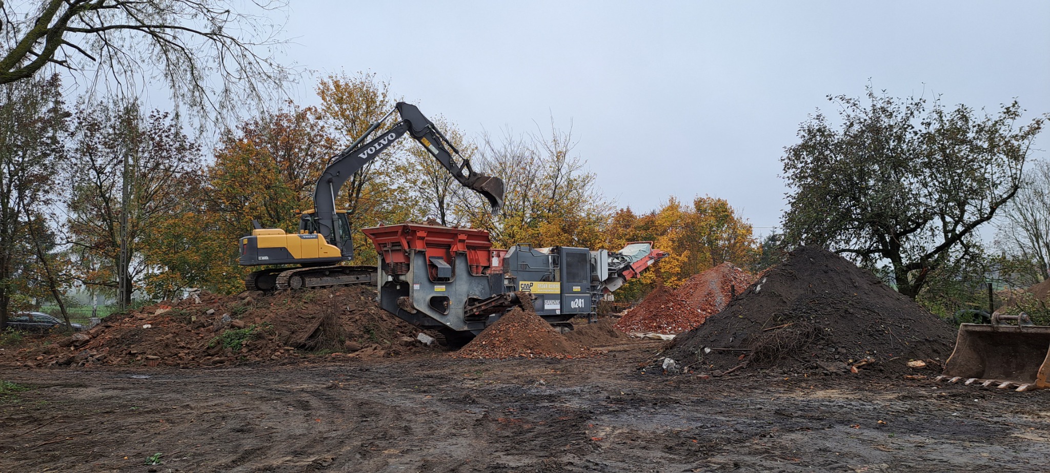 Żółta koparka Volvo załadowuje gruz do mobilnego przesiewacza Sandvik QJ341 na placu budowy z drzewami w tle.