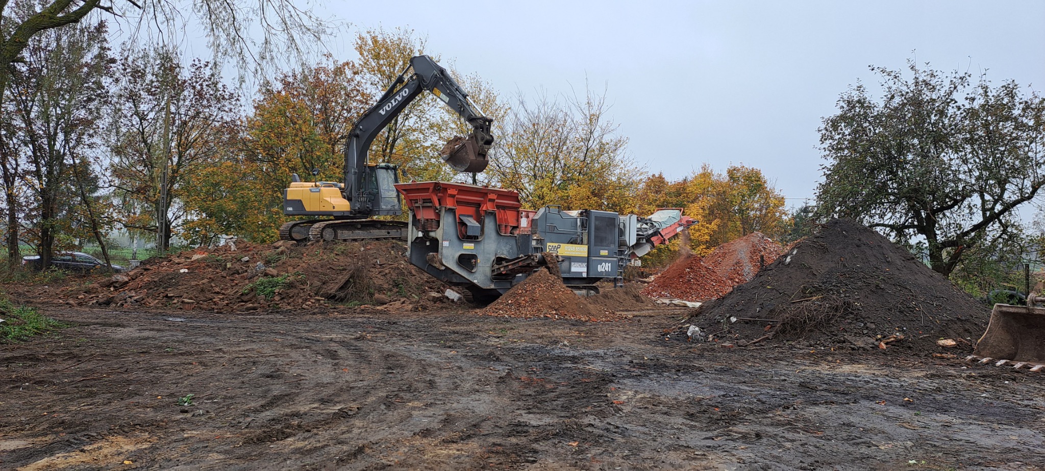 Żółta koparka Volvo umieszcza urobek w kruszarce mobilnej na tle jesiennych drzew, obok pryzmy gruzu i ziemi.