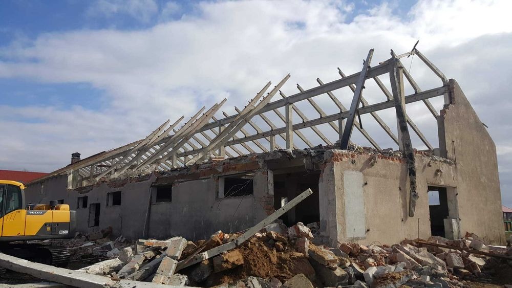 Częściowo zdemontowany budynek z odsłoniętą konstrukcją dachu, gruzem u podstawy i żółtą koparką Volvo w tle, ukazujący postęp prac rozbiórkowych.