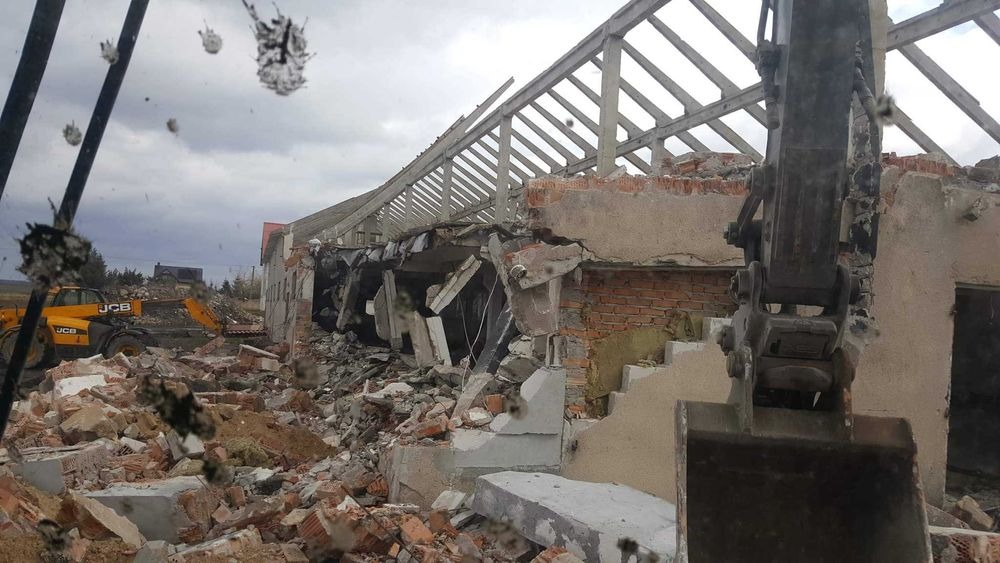 Częściowo zburzony budynek z widocznym szkieletem dachu, gruzem i koparką JCB w tle, ujęcie z wnętrza kabiny innej maszyny, z widocznymi zabrudzeniami na szybie.