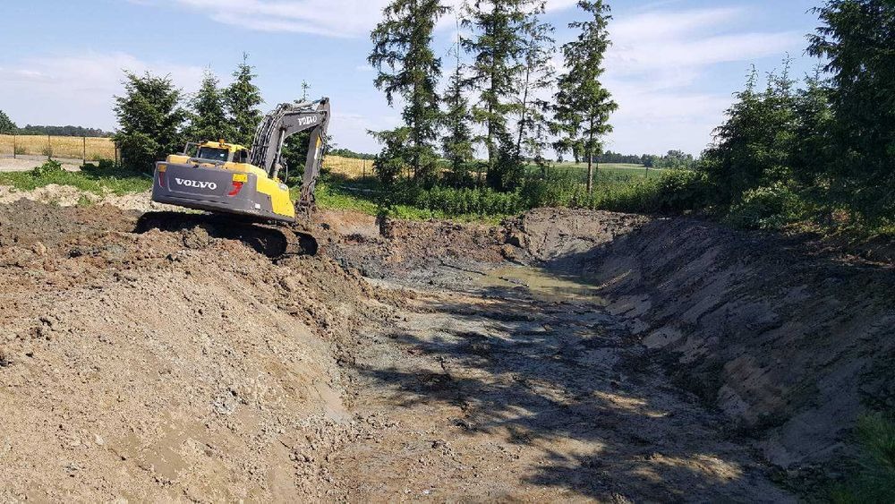 Żółto-szara koparka Volvo pracuje przy wykopie długiego rowu w ziemi, widok na teren budowy z drzewami w tle.