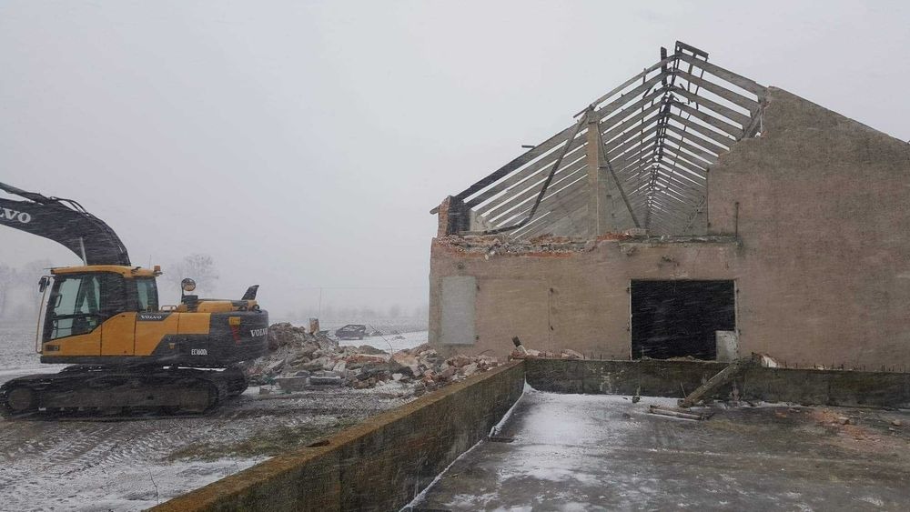 Żółta koparka Volvo EC160D w trakcie rozbiórki częściowo zburzonego budynku z widoczną drewnianą konstrukcją dachu podczas śnieżnej pogody.