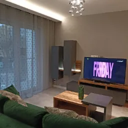 Nowoczesny salon z zieloną sofą, drewnianym stolikiem kawowym i telewizorem z napisem FRIDAY, oświetlony dekoracyjną lampą sufitową i listwą LED przy karniszu.