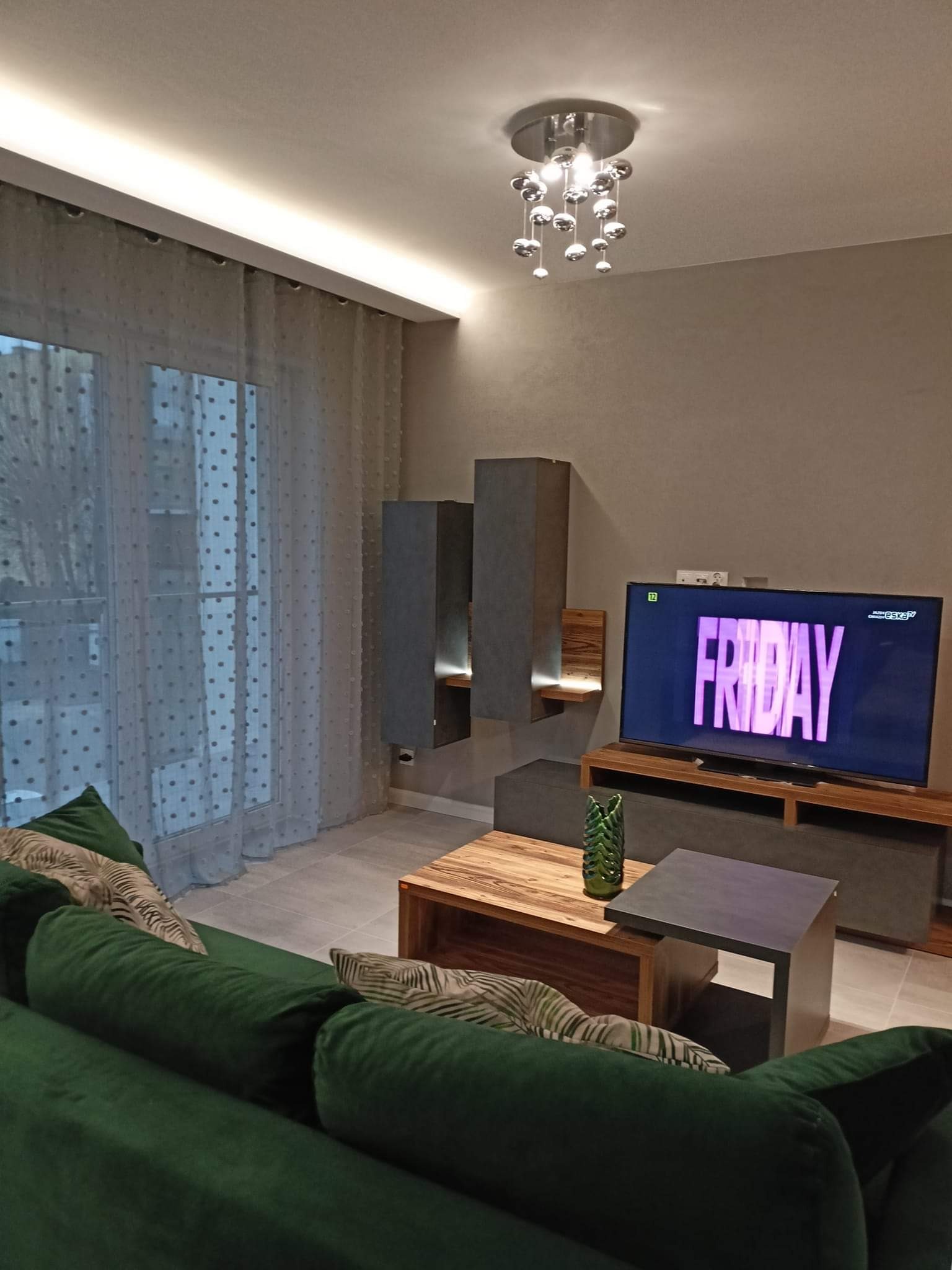 Nowoczesny salon z zieloną sofą, drewnianym stolikiem kawowym i telewizorem z napisem FRIDAY, oświetlony dekoracyjną lampą sufitową i listwą LED przy karniszu.
