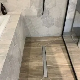 Wykończona łazienka z heksagonalnymi płytkami ściennymi w odcieniach szarości, podłogą imitującą drewno z liniowym odpływem prysznicowym i czarną ramą kabiny prysznicowej.