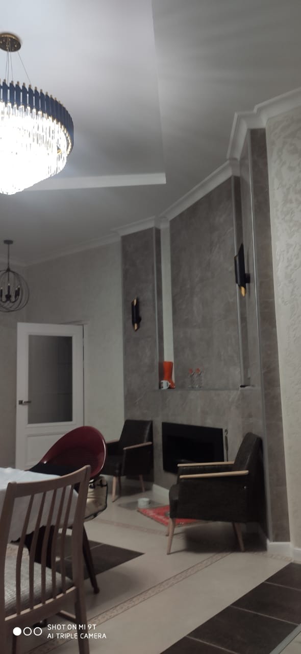 Jasne wnętrze pokoju z szarą ścianą akcentującą, ozdobioną listwami i dwoma czarnymi kinkietami, nad czarnym kominkiem z półką, na której stoi pomarańczowy but i szklanki, widoczne krzesła i duży...