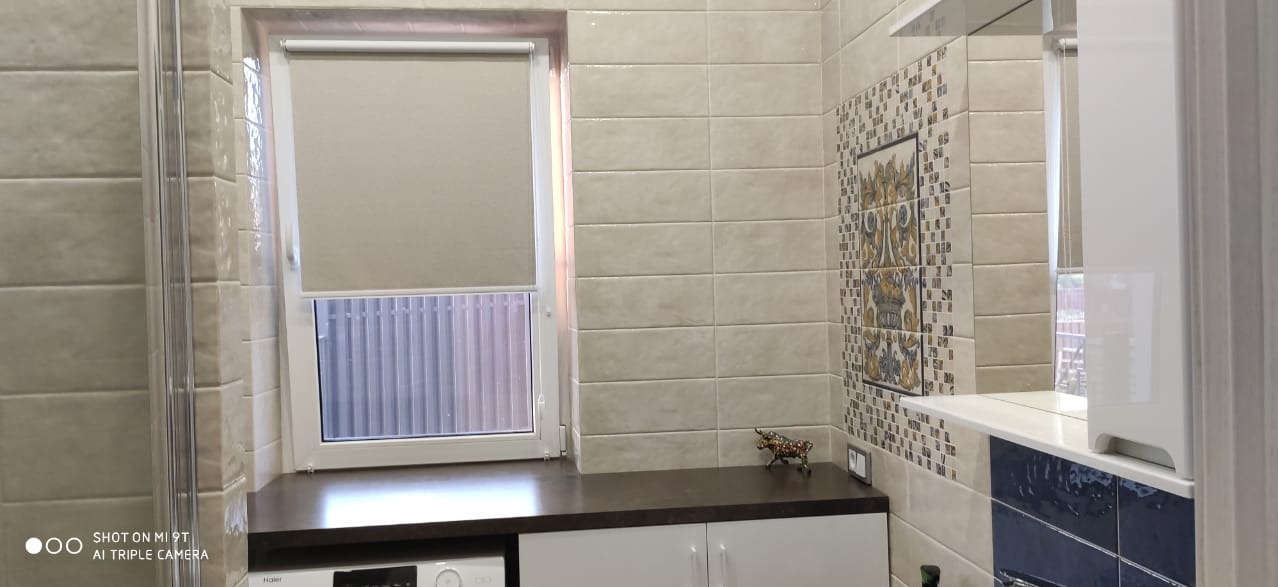 Wykończona łazienka z beżowymi płytkami imitującymi cegłę, dekoracyjną mozaiką, roletą w oknie i figurką byka na blacie.