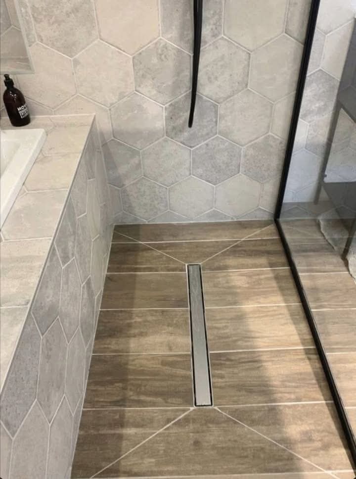 Wykończona łazienka z heksagonalnymi płytkami ściennymi w odcieniach szarości, podłogą imitującą drewno z liniowym odpływem prysznicowym i czarną ramą kabiny prysznicowej.