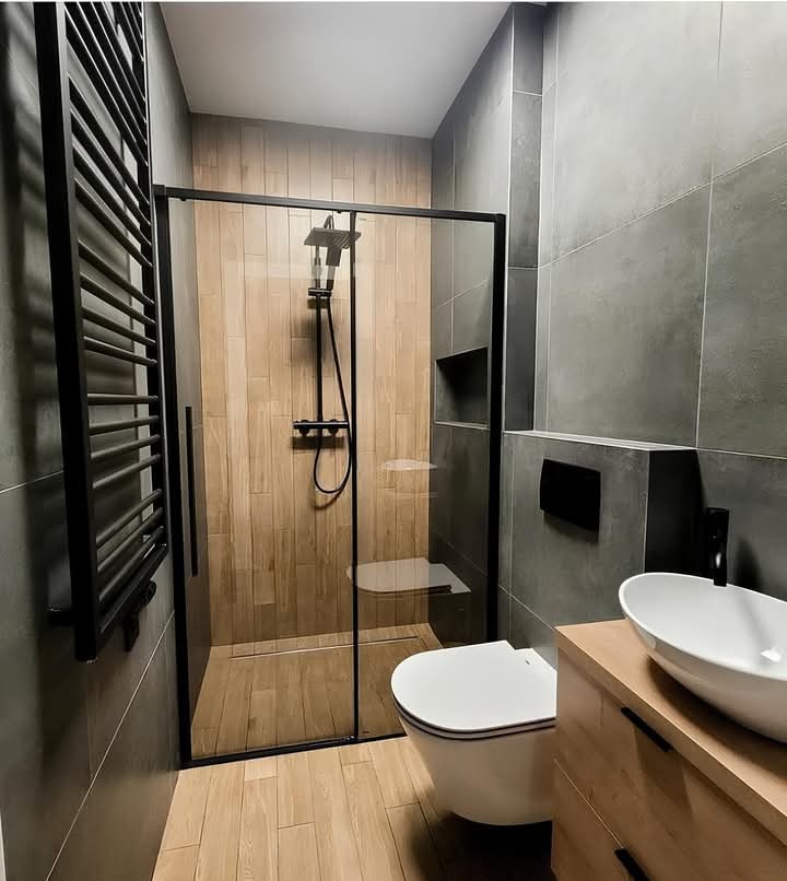 Nowoczesna łazienka z kabiną prysznicową typu walk-in z czarnymi profilami, grzejnikiem drabinkowym, podwieszaną toaletą i umywalką nablatową na drewnianej szafce, wykończona szarymi płytkami...