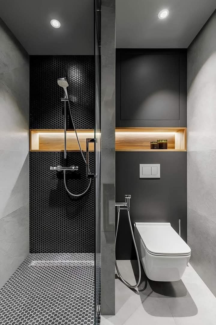 Nowoczesna łazienka w odcieniach szarości z czarną mozaiką heksagonalną pod prysznicem i drewnianymi akcentami w podświetlonych wnękach, minimalistyczna toaleta podwieszana z bidetką.