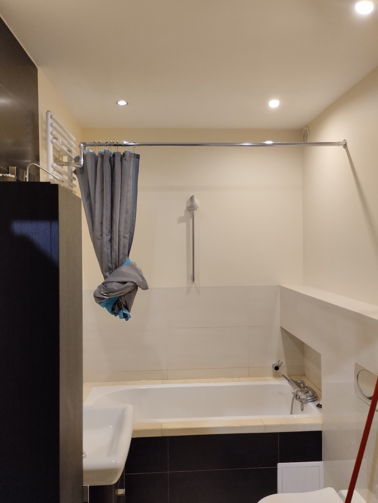 Nowoczesna łazienka z wanną, szarą zasłoną prysznicową z akcentami turkusowymi i czarnymi płytkami pod wanną, oświetlona trzema punktowymi lampami.