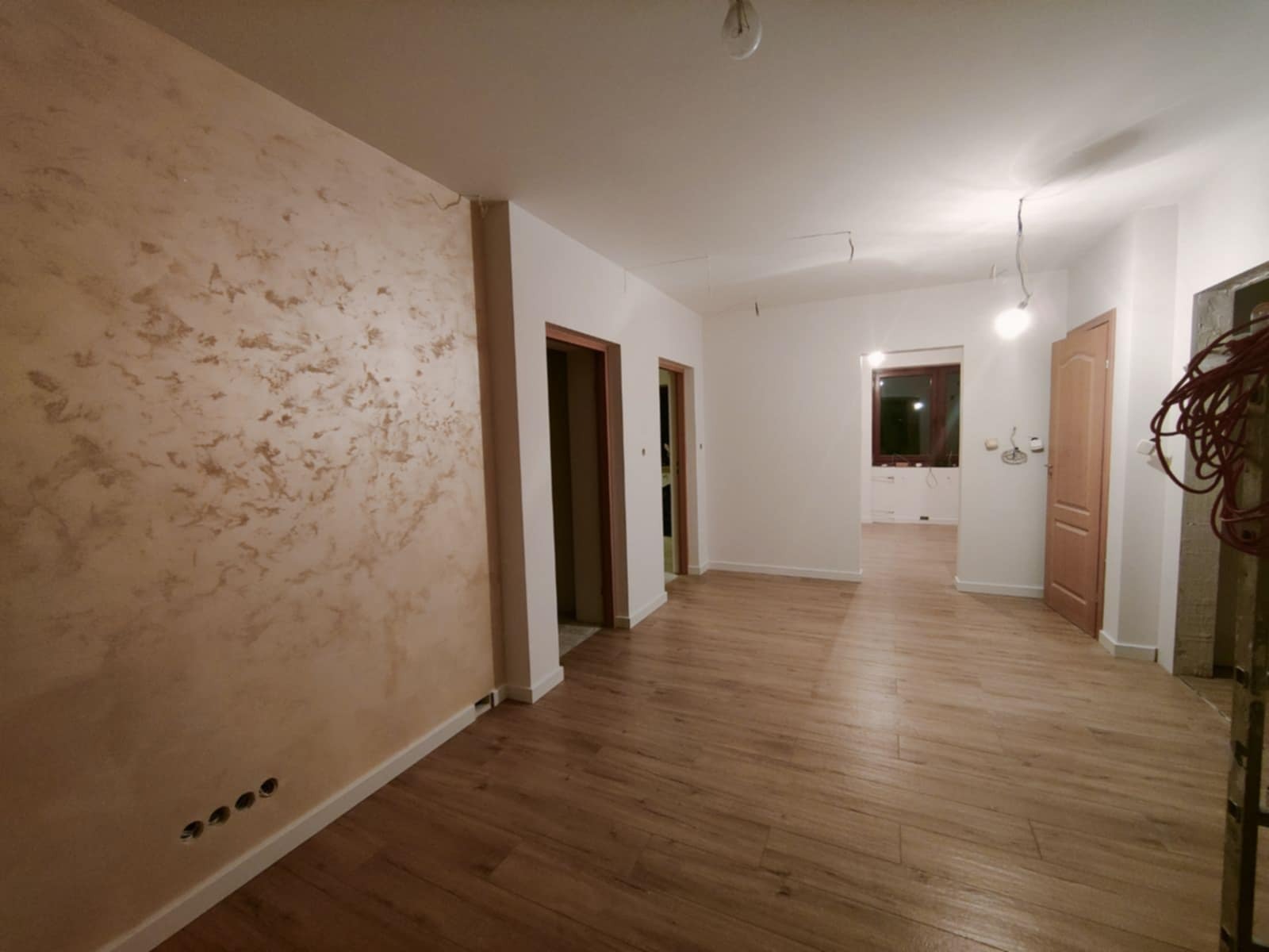 Pokój po remoncie z dekoracyjną ścianą w odcieniach beżu i brązu, drewnopodobną podłogą, widocznymi gniazdkami elektrycznymi i otwartymi przejściami do innych pomieszczeń.