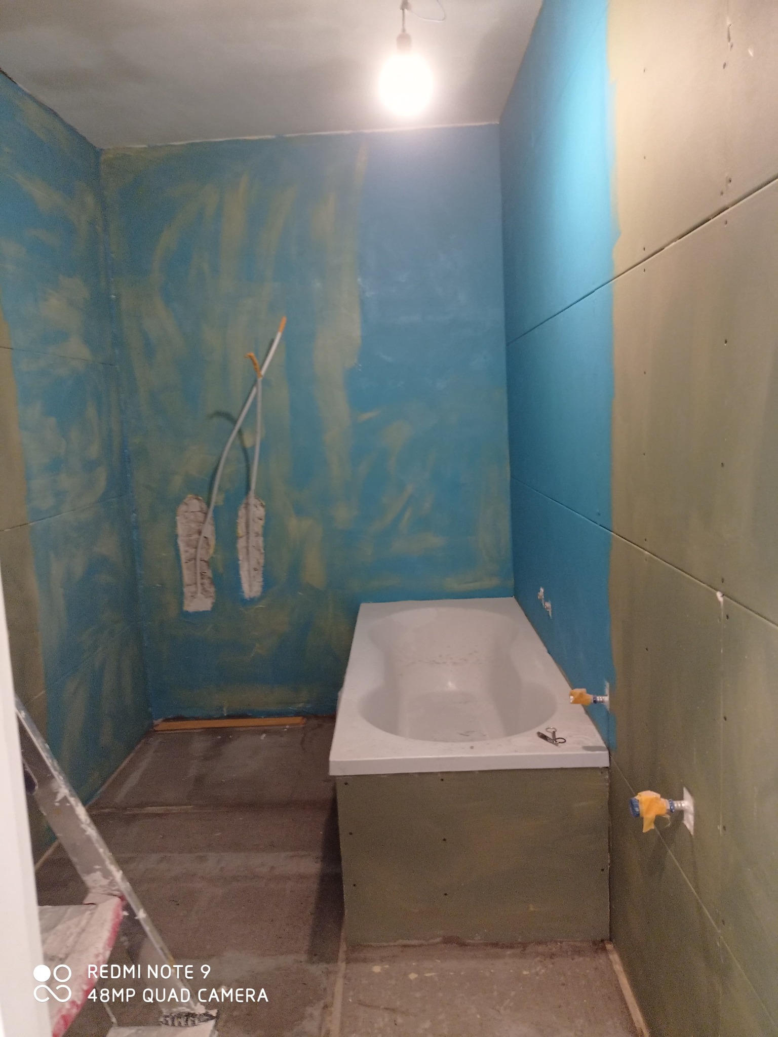 Łazienka w trakcie remontu: ściany częściowo pokryte niebieską farbą, częściowo płytami kartonowo-gipsowymi, widoczna wanna, rury z zabezpieczeniami, drabina i narzędzia.