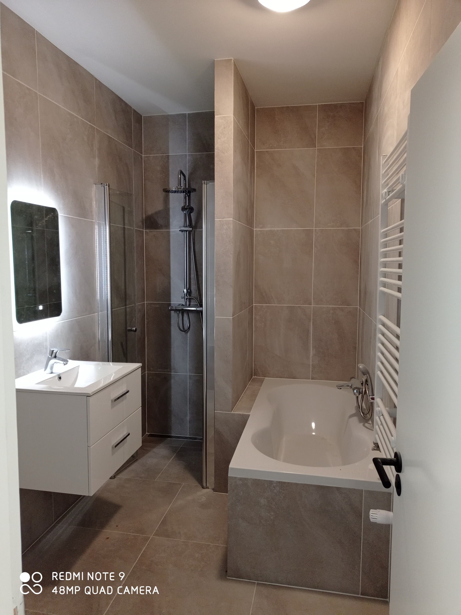 Nowoczesna łazienka z beżowymi płytkami, białą wiszącą szafką z umywalką, kabiną prysznicową z czarną armaturą oraz wanną z grzejnikiem drabinkowym.