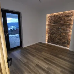 Wykończone wnętrze pokoju z drewnianą podłogą, ścianą z cegieł podświetloną od tyłu oraz widokiem na ośnieżone g&oacute;ry przez przeszklone drzwi balkonowe.