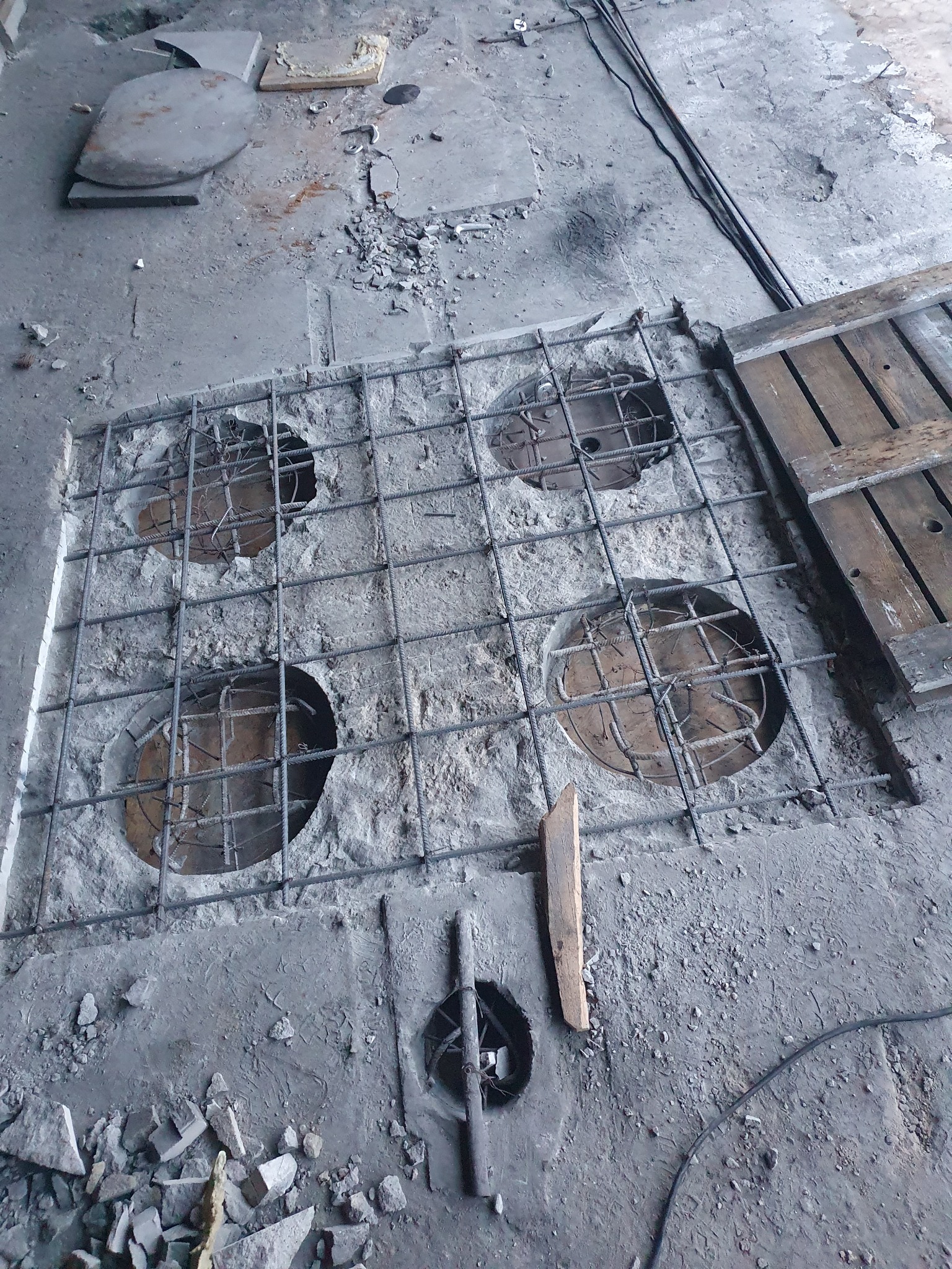 Betonowy strop z wyciętymi okrągłymi otworami, zbrojony siatką stalową, widoczne fragmenty gruzu i desek szalunkowych.