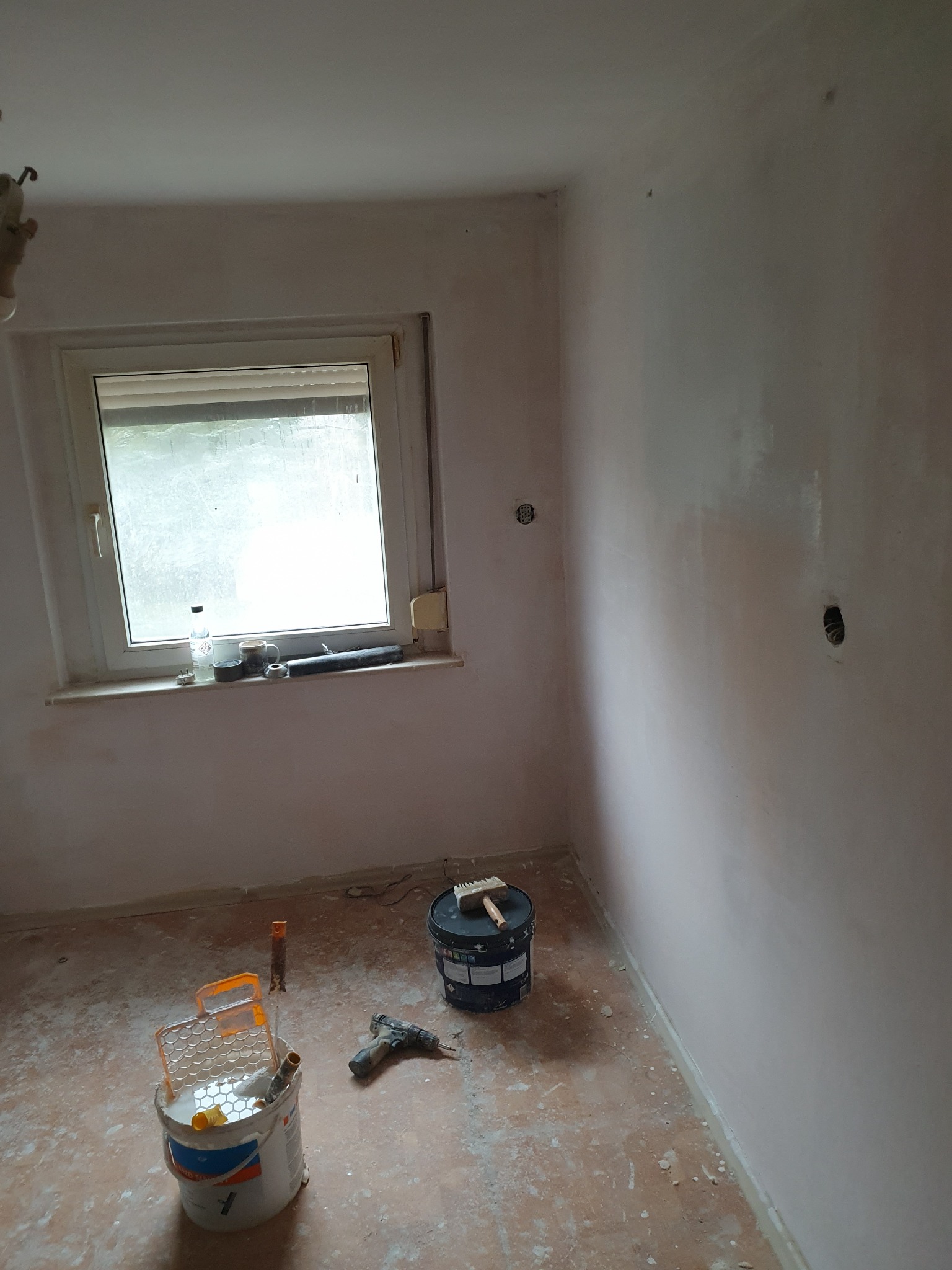 Pokój w trakcie odświeżania: ściany po nałożeniu pierwszej warstwy farby, wiadro z farbą, wałek malarski i wiertarka na podłodze, okno z przedmiotami na parapecie.